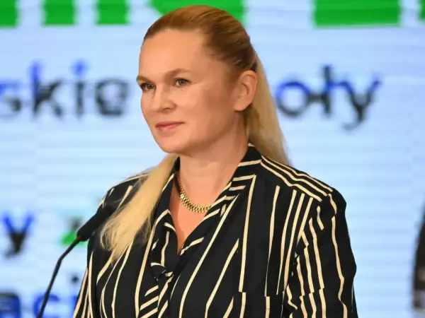 Kobieta z blond włosami w pasiastym ubraniu stoi przed mikrofonem na tle ekranu z tekstem.