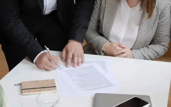 Dwie osoby przy stole podpisują dokumenty. Jedna z nich pisze piórem.