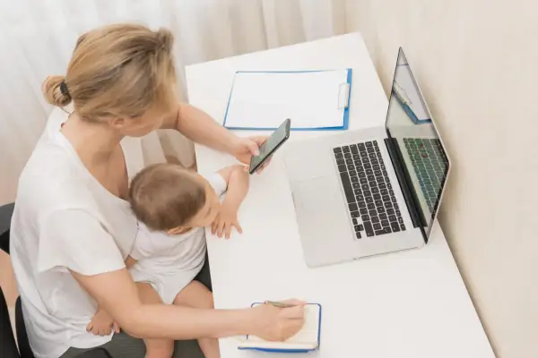 Kobieta z dzieckiem na kolanach sprawdza na telefonie, urlop macierzyński ile płatny. Obok laptop i notatnik.