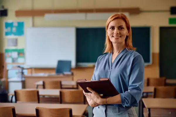 Uśmiechnięta nauczycielka z teczką w ręku stoi przed klasą z ławkami i tablicą.
