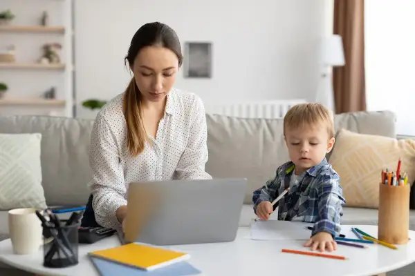Kobieta pracuje przy laptopie, podczas gdy jej syn rysuje obok. To obrazek z życia, gdzie praca i rodzina łączą się, jak w przypadku art. 188 kodeksu pracy.