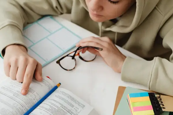 Osoba w bluzie z kapturem przegląda książkę, obok leżą okulary i kolorowe karteczki.