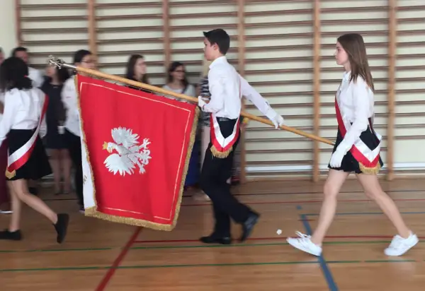 Uczniowie w galowych strojach tworzą poczet sztandarowy, niosąc biało-czerwony sztandar z godłem Polski.