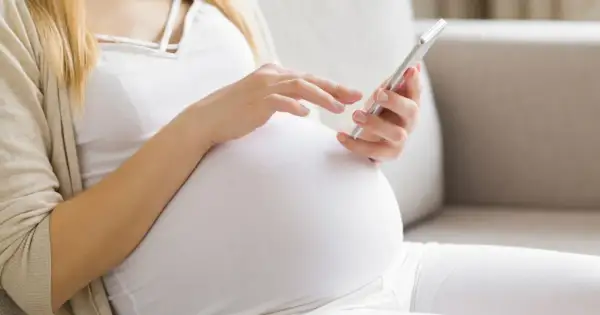 Ciężarna kobieta w białej bluzce i beżowym kardiganie używa telefonu komórkowego.
