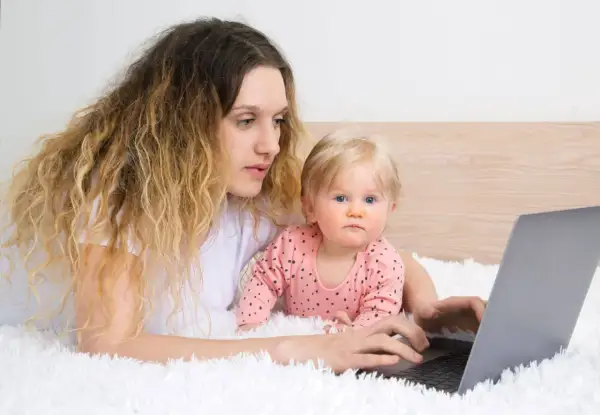 Kobieta z dzieckiem na rękach pracuje przy laptopie, zastanawiając się, czy na urlopie wychowawczym można pracować.